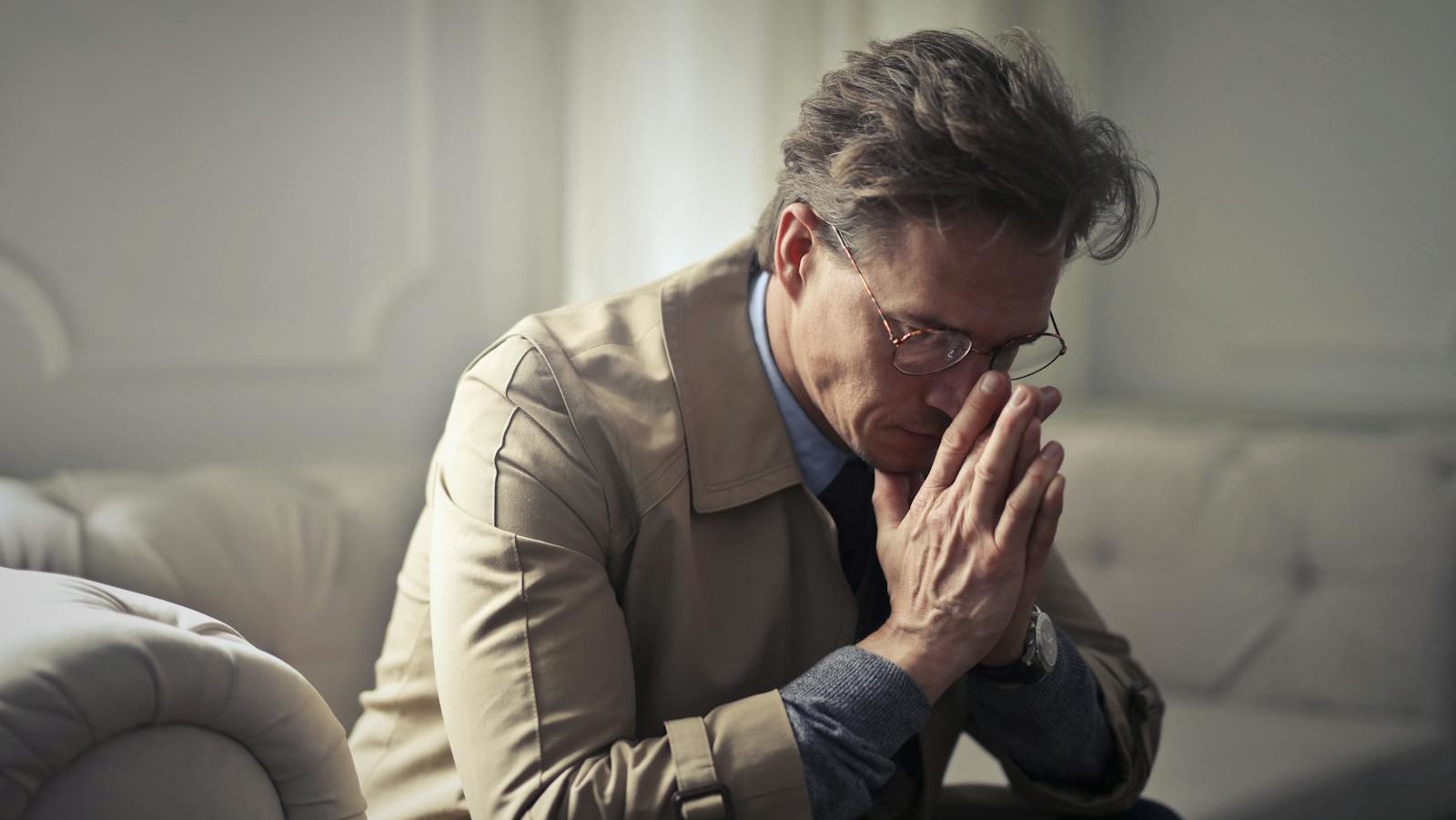 Side view of upset male entrepreneur in formal wear sitting on couch in modern living room and contemplating about financial problems while leaning on hands and looking down