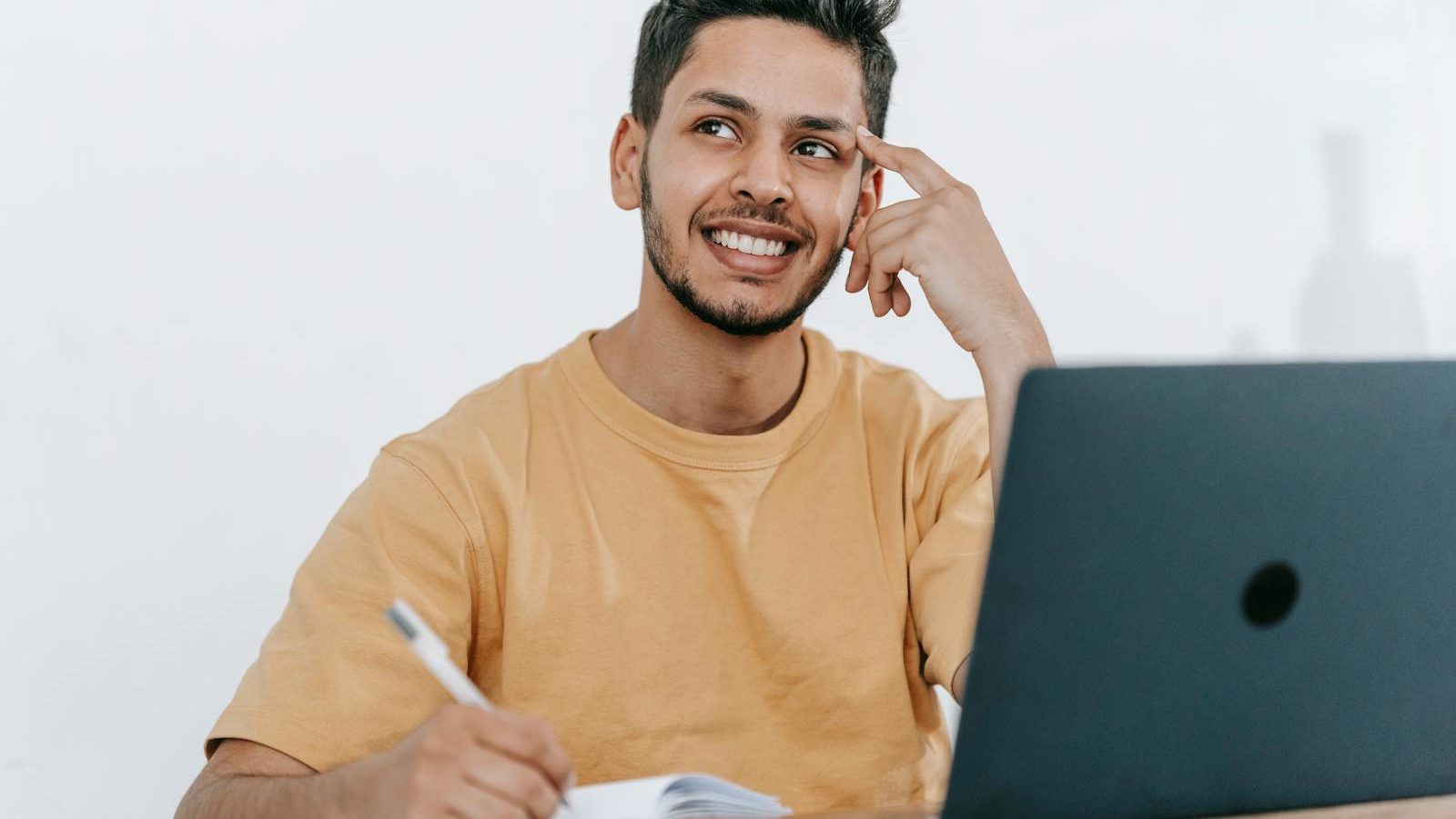 Smiling young bearded Hispanic male entrepreneur thinking over new ideas for startup project and looking away dreamily while working at table with laptop and taking notes in notebook