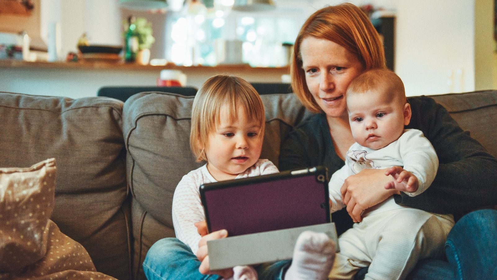 two babies and woman sitting on sofa while holding baby and watching on tablet